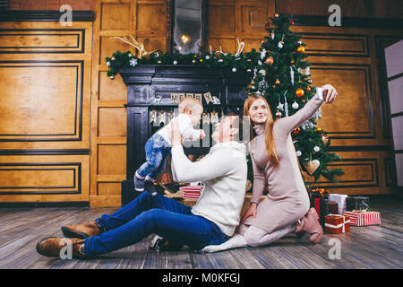 Tema Natale. Il Padre genera il figlio del biondino di un anno di età, sedersi su una struttura in legno nella parte anteriore di albero di Natale con doni, mom rende selfie, self-portr Foto Stock