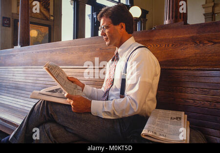 Un " commuter " legge un giornale finanziario in una stazione ferroviaria come attende. Foto Stock