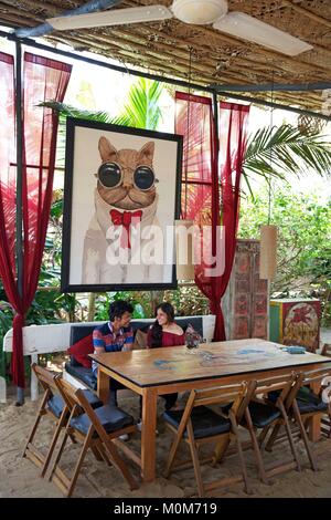 L'India,Goa,Morjim beach,coppia giovane posti a sedere sotto un dipinto ritratto di gatto nel ristorante sulla spiaggia la Plage Foto Stock