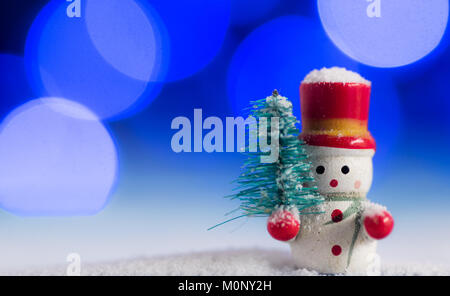 Carino pupazzo di neve decorazione di Natale, con splendidi blu e viola bokeh sfondo luci, sulla sommità del bianco della neve, tenendo un pino Foto Stock