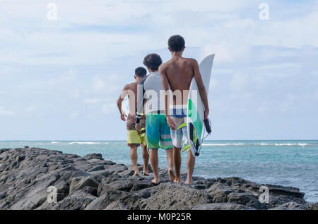 Tre ragazzi adolescenti a piedi lungo la parete di roccia a waterfront. Surfisti di adolescenti a piedi per prendere le onde. Ragazzi a piedi per la ricerca di un'onda - Hawaii, STATI UNITI D'AMERICA Foto Stock