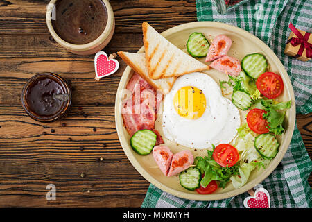 La prima colazione il giorno di San Valentino - uovo fritto a forma di cuore, toast, salsicce, pancetta e verdure fresche. Prima colazione inglese. Tazza di caffè. Top v Foto Stock