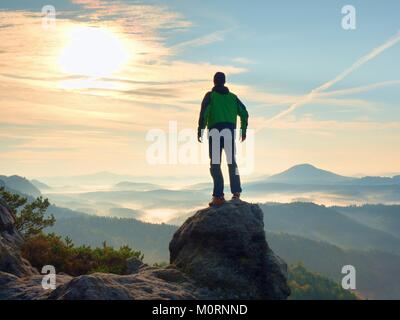 L'uomo rimanere sul picco roccioso entro lo spuntar del giorno e guardare oltre il brumoso paesaggio. Bella giornata autunnale. Foto Stock