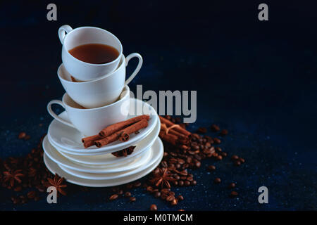 La testata con una pila di bilanciamento del bianco tazze di caffè e tè piattini, i chicchi di caffè e cannella su uno sfondo blu scuro. Dark cibo fotografia con co Foto Stock