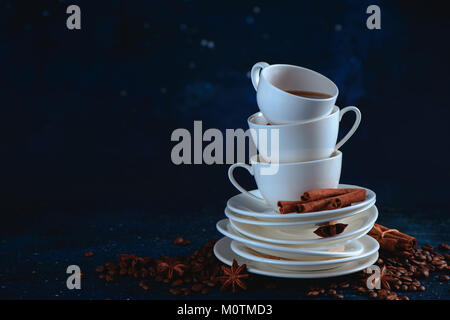 La testata con una pila di bilanciamento del bianco tazze di caffè e tè piattini, i chicchi di caffè e cannella su uno sfondo blu scuro. Dark cibo fotografia con co Foto Stock