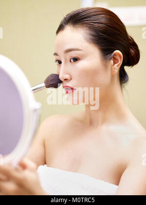 Bella giovane donna asiatica guardando nello specchio mentre si applica il make-up sul viso utilizzando una spazzola. Foto Stock