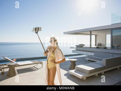 Donna in costume da bagno tenendo selfie con selfie stick sulla soleggiata, moderno luxury home vetrina patio esterno con vista oceano Foto Stock