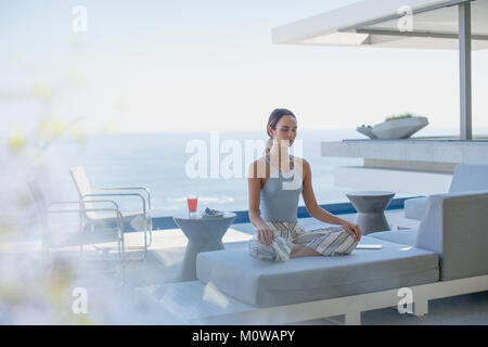 Serena donna meditando nella posizione del loto sul moderno, lusso home vetrina patio esterno divano con vista oceano Foto Stock