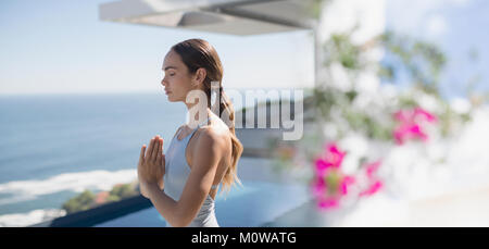 Serena brunette donna a praticare yoga e meditazione con le mani al centro del petto sul patio soleggiato con vista oceano Foto Stock