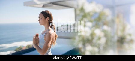 Serena brunette donna a praticare yoga e meditazione con le mani al centro del petto sul patio soleggiato con vista oceano Foto Stock