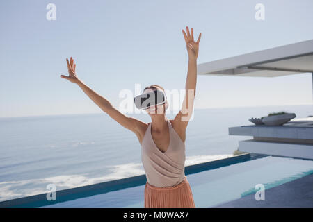 Donna energica utilizzando la realtà virtuale occhiali simulatore sulle moderne, lusso home vetrina patio esterno con vista oceano Foto Stock