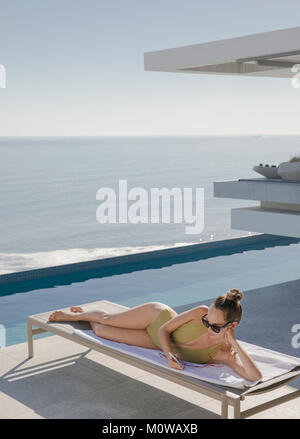 La donna a prendere il sole, sms con smart phone su sedia a sdraio sul soleggiato patio di lusso con vista oceano Foto Stock