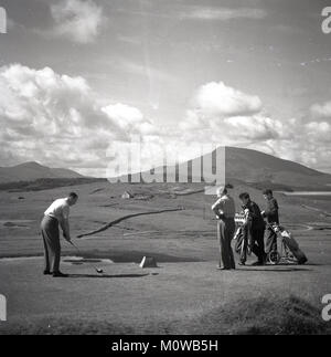 Degli anni Cinquanta, storico, un golfista maschio guida fuori da un tee off su un entroterra aperto campo da golf in Irlanda, con il suo modo di suonare il partner e i loro due giovane ragazzo caddie guardando su. Foto Stock