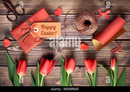 Dono avvolto con tag 'Felice Pasqua', materiali di imballaggio, forbici e tulipani rossi su un tavolo di legno. Concetto di preparazione alle vacanze di Pasqua testo spac Foto Stock
