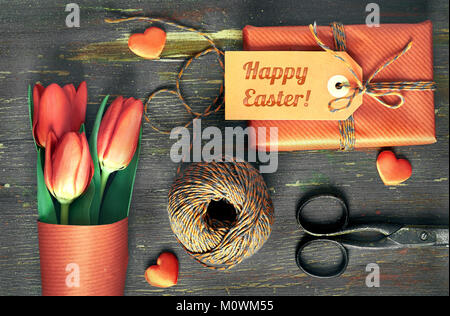 Dono avvolto con tag 'Felice Pasqua', materiali di imballaggio, forbici e tulipani rossi su un tavolo di legno. Concetto di preparazione alle vacanze di Pasqua. Foto Stock