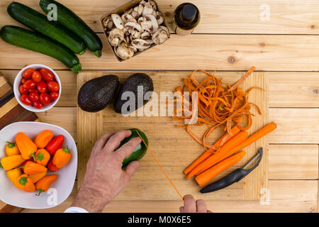 Chef per affettare verdura fresca per la cottura su un tagliere di legno tenendo un peperone verde con carote pelate, zucchine, funghi, pomodori,mature av Foto Stock