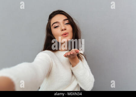 Close-up verticale di gioiosa brunette donna di inviare aria kiss tenendo selfie sul telefono cellulare, isolate su uno sfondo grigio Foto Stock