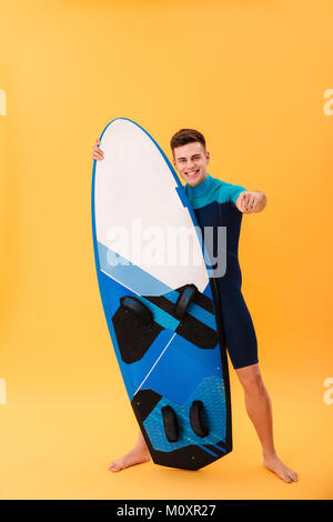 A piena lunghezza Ritratto di fiducioso uomo sorridente in costume da bagno puntando con il dito su di voi mentre in piedi e tenendo le tavole da surf, isolato su giallo backg Foto Stock