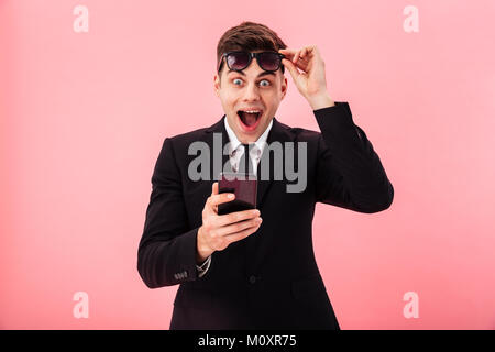 Sorpreso uomo felice decollare occhiali da sole utilizza lo smartphone isolato su rosa Foto Stock