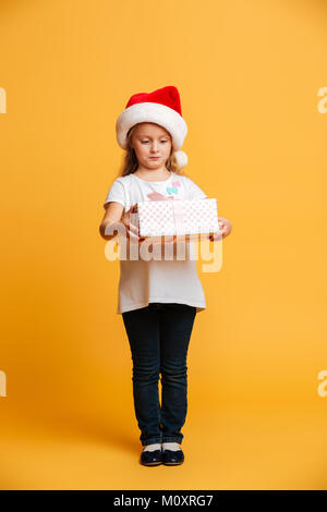 Immagine della piccola ragazza concentrato in piedi isolato su sfondo giallo che indossa christmas santa hat. Guardando oltre tenendo sorpresa confezione regalo. Foto Stock