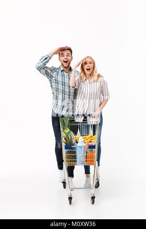 A piena lunghezza Ritratto di un allegro giovane in piedi con un carrello per supermercati isolate su sfondo bianco Foto Stock