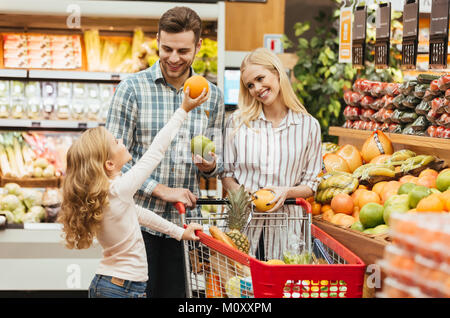 Felice famiglia giovane in piedi con un carrello e scegliendo la frutta al supermercato Foto Stock