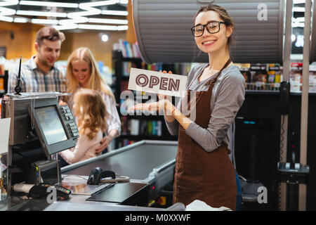 Ritratto di donna felice cassiere tenendo aperto mentre il segno permanente al contatore di cassa nel supermercato Foto Stock