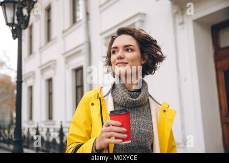 Foto di allegro piuttosto giovane donna vestita in giallo impermeabile passeggiate all'aperto a bere caffè. Guardando a parte. Foto Stock