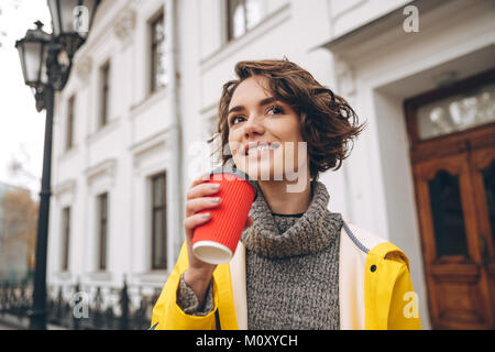 Foto di allegro piuttosto giovane donna vestita in giallo impermeabile passeggiate all'aperto a bere caffè. Guardando a parte. Foto Stock