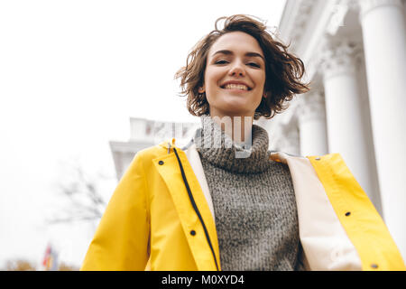 Closeup ritratto di incantevole giovane donna con bob haircut godendo a piedi attraverso la città nel rivestimento giallo sorridente su telecamera Foto Stock