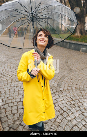 Bella lieta adulto ragazza in giallo impermeabile in piedi sotto il grande ombrello trasparente, con un largo sorriso sincero nel giardino della città Foto Stock