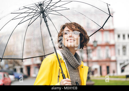 Splendido ritratto di donna felice in giallo impermeabile a camminare in città sotto ombrello trasparente, durante il freddo giorno di pioggia Foto Stock