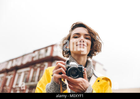 Foto della splendida signora giovane fotografo vestito con un impermeabile all'aperto a piedi tenendo la fotocamera. Foto Stock