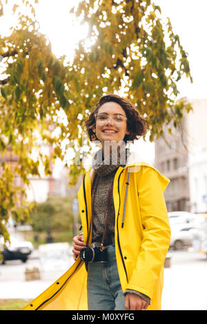 Immagine di allegra donna giovane fotografo vestito con un impermeabile all'aperto a piedi con la fotocamera. Foto Stock