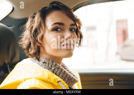 Immagine di piuttosto giovane signora vestita di impermeabile seduti in auto. Guardando la fotocamera. Foto Stock