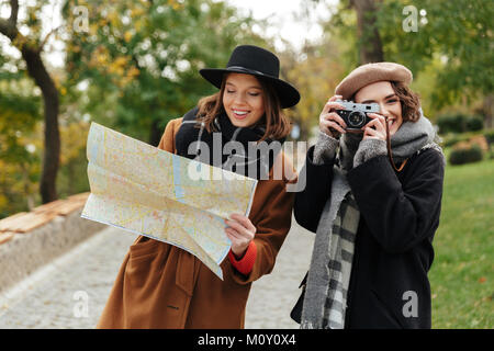 Ritratto di due attraenti ragazze vestiti in abiti d'autunno azienda mappa della città mentre in piedi all'aperto e fare le foto con la fotocamera vintage Foto Stock