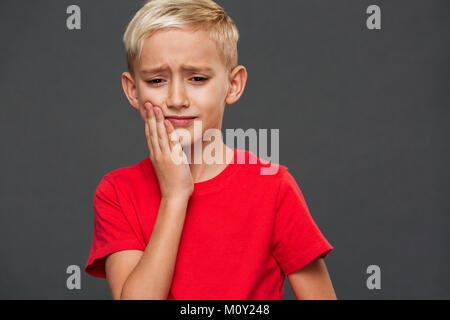 Immagine della sad little boy bambino con il mal di denti in piedi isolato su sfondo grigio. Guardando a parte. Foto Stock