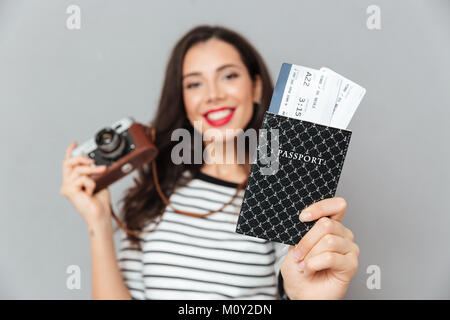 Ritratto di una giovane donna tenendo la fotocamera vintage e mostrando passaporto con i biglietti di volo isolate su uno sfondo grigio Foto Stock