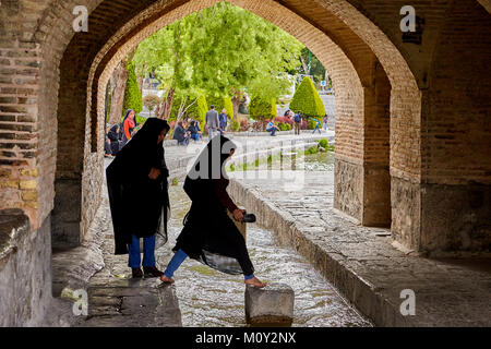 Isfahan, Iran - 24 Aprile 2017: due giovani iraniane di donne musulmane, vestito di un nero hijab islamico, saltando su di un flusso di acqua sotto il ponte alla Foto Stock