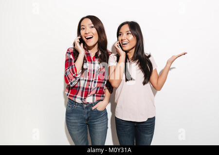 Immagine di donne asiatiche sorelle parlando dai loro telefoni cellulari isolate in piedi oltre il muro bianco sullo sfondo. Foto Stock