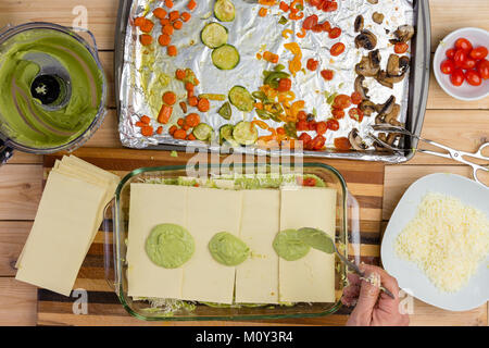 L'uomo preparare una lasagna con verdure fresche spooning pere di avocado su uno strato di pasta essiccata in un forno per vetro piatto per la cottura visto dal di sopra Foto Stock