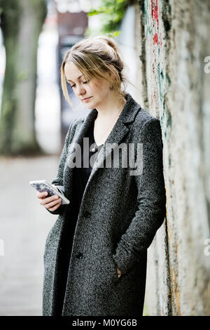 Giovane donna si appoggia contro un muro e guarda al suo smartphone,di Colonia, nella Renania settentrionale-Vestfalia,Germania Foto Stock