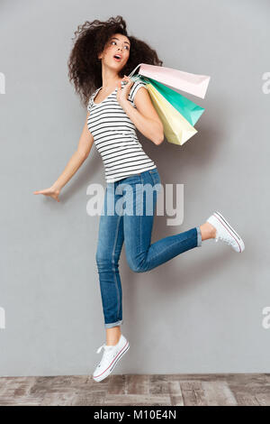 Caucasian brunette femmina nel casual in posa sulla fotocamera con un sacco di pacchetti in mano, essendo soddisfatto dopo lo shopping oltre il muro grigio in studio Foto Stock