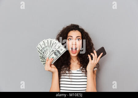 Lucky donna con capelli ricci mantenendo la ventola di 100 fatture del dollaro e smartphone in mani che mostra è possibile guadagnare un sacco di soldi usando gadget elettronici Foto Stock