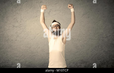 Un giovane uomo con la barba, headstrap e bicchieri in posa di fronte dello sbozzato muro grigio Sfondo, immaginando ha grandi muscoli Foto Stock