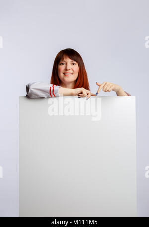 Foto di bruna chef in veste bianca con vuoto poster su sfondo bianco in studio Foto Stock