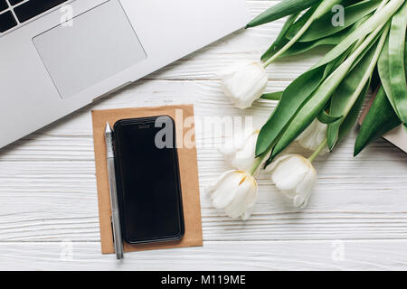 Lo schermo del telefono e computer portatile e tulipani su legno bianco sfondo rustico. elegante laici piatto con fiori e gadget di lavoro con spazio per il testo. freelance Foto Stock