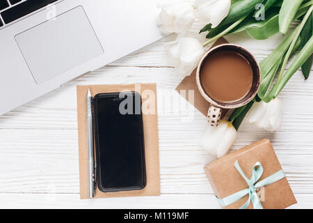 Lo schermo del telefono e computer portatile con il caffè del mattino e tulipani su legno bianco sfondo rustico. elegante laici piatto con fiori e gadget di lavoro con spazio Foto Stock