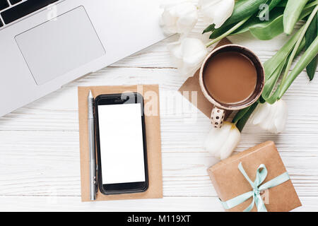 Svuotare lo schermo del telefono e computer portatile con il caffè del mattino e tulipani su legno bianco sfondo rustico. elegante laici piatto con fiori e gadget di lavoro con Foto Stock