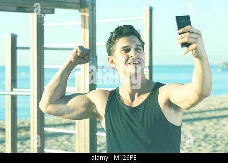 Allegro uomo sportivo che mostra e bicipiti tenendo selfie sulla spiaggia stazione fitness Foto Stock